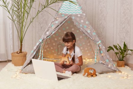 Photo for Indoor shot of satisfied delighted little girl with pigtails lying in peetee tent with laptop and having online conversation with her friend, showing her new wooden horse. - Royalty Free Image