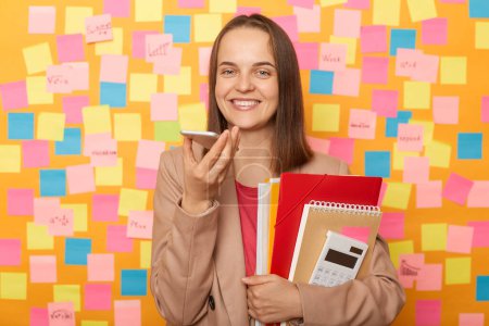Téléchargez les photos : Image of charming cute beautiful woman wearing beige jacket, holding her documents, recording voice message or commands standing against yellow wall with colorful stickers. - en image libre de droit