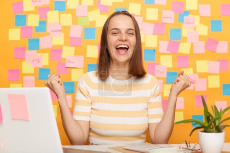Foto de Horizontal shot of excited amazed happy young adult woman wearing T-shirt posing against yellow paper wall with adhesive notes, clenched fists, celebrating her victory. - Imagen libre de derechos
