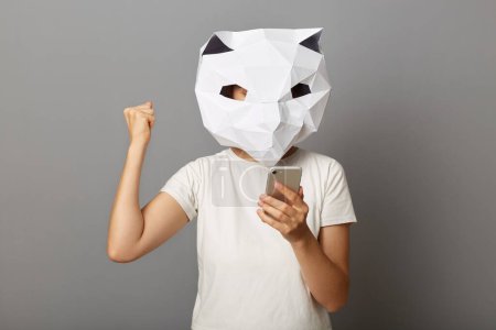 Téléchargez les photos : Photo d'une joyeuse femme joyeuse portant un masque en papier pour chat et un T-shirt blanc, utilisant un téléphone portable, un poing serré, célébrant la victoire, posant isolée sur un fond gris. - en image libre de droit