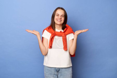 Photo for Image of cheerful joyful female with brown hair wearing white T-shirt and orange jumper tied over shoulders, standing isolated on blue background, standing with raised arms, shrugging shoulders. - Royalty Free Image