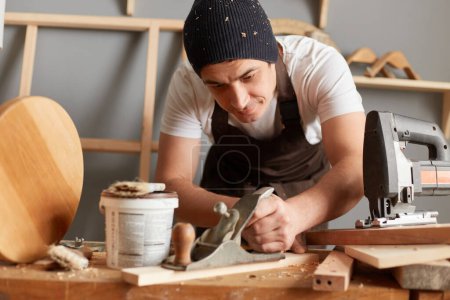 Photo for Indoor shot of concentrated man carpenter wearing apron and cap working with plane on wooden workplace background, making wooden furniture, enjoying his work in workshop, handmade wooden products. - Royalty Free Image