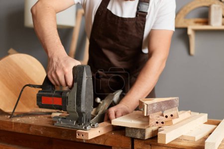 Photo for Portrait of faceless anonymous carpenter working on woodworking machines in carpentry shop, holding jigsaw in hands, standing at workbench, using instrument. - Royalty Free Image