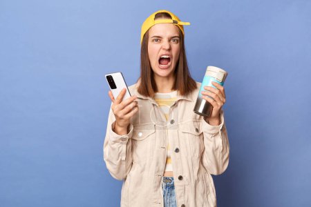 Photo for Portrait of angry aggressive teenager girl wearing baseball cap and jacket, holding thermos, using cell phone, expressing hate and anger, screaming, posing isolated over blue background. - Royalty Free Image