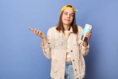 Photo for Portrait of confused uncertain teenager girl wearing baseball cap and jacket posing isolated over blue background, holding mobile phone, dosen't know what to answer, reading message. - Royalty Free Image