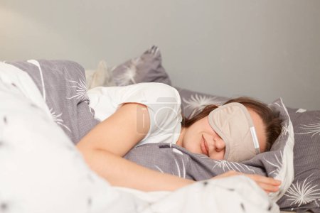 Foto de Indoor shot of young woman wearing sleepwear sleeping in a bed with eyes covered with gray mask, lying under blanket, can sleep longer during weekend. - Imagen libre de derechos