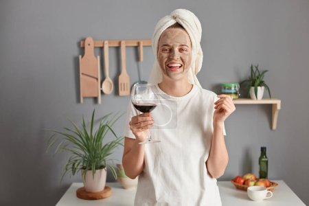 Foto de Image of joyful attractive winsome woman with cosmetic mask in white towel wrapped around head drinking wine in kitchen, rejoicing her day off, celebrating happy weekend, looking at camera. - Imagen libre de derechos