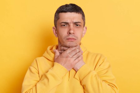 Photo for Portrait of ill man with dark hair wearing casual style hoodie standing isolated over yellow background, touching his neck, suffering sore throat, viral infection or flu symptoms. - Royalty Free Image
