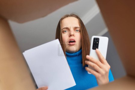 Photo for Shocked sad woman received parcel with wrong or damages items, holding waybill in hands and mobile phone, calling to service, looking inside of box with astonished expression. - Royalty Free Image