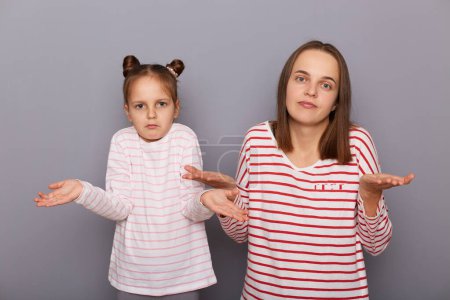 Photo for Portrait of uncertain woman and little girl with hair buns wearing casual clothes standing isolated over gray background, family posing with shrugging shoulders, having doubtful expression. - Royalty Free Image