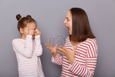 Téléchargez les photos : Indoor shot of aggressive angry woman and crying little girl with hair buns wearing casual clothes standing isolated over gray background, mother scolds daughter for bad grades and behavior. - en image libre de droit
