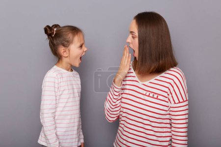 Téléchargez les photos : Indoor shot of shocked amazed mother and screaming daughter wearing casual clothes standing isolated over gray background, small kid yelling loud, mother with surprised face, covering mouth with hand. - en image libre de droit