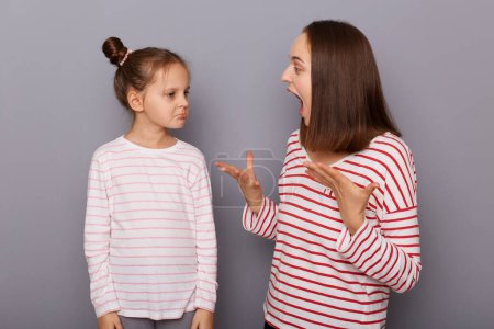 Téléchargez les photos : Portrait of irritated angry mother and sad daughter wearing casual clothes standing isolated over gray background, woman arguing with her kid, screaming with raised arms. - en image libre de droit