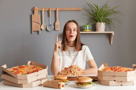 Photo for Portrait of inspired amazed excited woman with brown hair wearing white T-shirt sitting at table, being surrounded with junk food, raised finger up, has idea. - Royalty Free Image