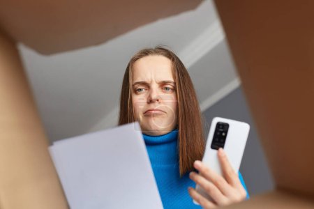 Photo for Sad serious woman customer wearing blue turtleneck opening parcel, looking at consignment, trying to understand where is a mistake, holding phone, needs to call to delivery service. - Royalty Free Image