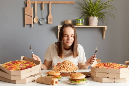 Photo for Horizontal shot of satisfied woman with brown hair wearing white casual T-shirt sitting at table in kitchen, posing with closed eyes, smelling pasta, pizza, hamburgers. - Royalty Free Image