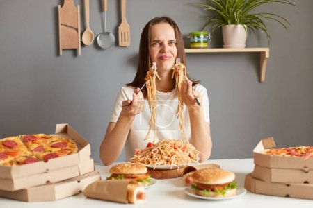Photo for What an awful meal? Irritated frustrated woman with brown hair wearing white casual T-shirt sitting at table in kitchen, looking at pasta and other fast food, having shocked expression. - Royalty Free Image