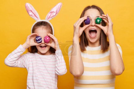 Téléchargez les photos : Photo d'une mère et d'une fille positives et joyeuses dans des oreilles de lapin couvrant les yeux avec des œufs de Pâques, s'amusant ensemble, célébrant les vacances en famille, se tenant isolées sur fond jaune - en image libre de droit