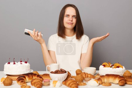 Photo for Portrait of uncertain woman with brown hair sitting at table with smart phone in hands, ordering lots sugary desserts, do not know how to eat all isolated over gray background. - Royalty Free Image