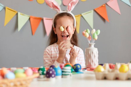 Portrait of happy funny little girl wearing rabbit ears sitting at table having fun while preparing for Easter, covering eyes with candy pops, laughing, against gray wall with decorations.