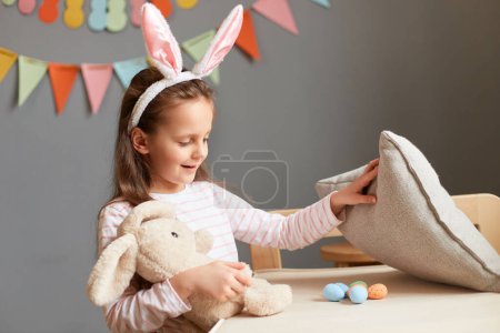 Photo of smiling happy cheerful little girl wearing rabbit ears finds eggs under pillow Easter hunt posing in home interior with decorations, celebrating holiday, playing.