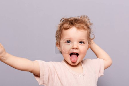 Cheerful smile child. Indoor shot of little infant girl showing tongue, standing with wide open mouth and sticking tongue out isolated over gray background.