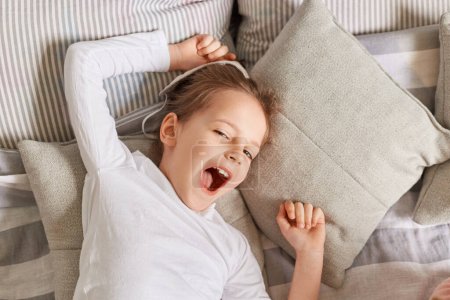 Photo for Sleepy little girl yawning in the morning while lying in bed on pillows, keeps mouth open lazy morning on weekend stretching arms with happy facial expression. - Royalty Free Image