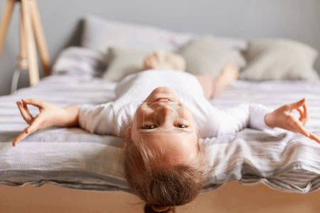 Photo for Happy little girl with dark hair lying upside down on bed at home, kid playing having fun showing victory looking smiling to camera. - Royalty Free Image