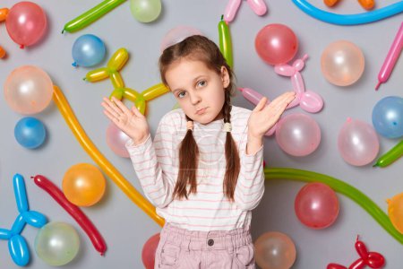 Photo for Uncertain confused little girl with pigtails standing against gray wall with balloons decoration shrugging shoulders looking at camera with puzzled facial expression. - Royalty Free Image