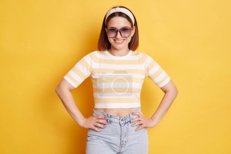 Photo for Confident cheerful woman wearing striped T-shirt hair band and sunglasses standing isolated over yellow background posing with hands on hips being in good mood. - Royalty Free Image