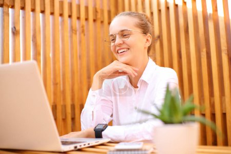 Satisfied smiling woman wearing white shirt sitting in outdoor cafe working on laptop having online meeting video conferencetalking with business partner.