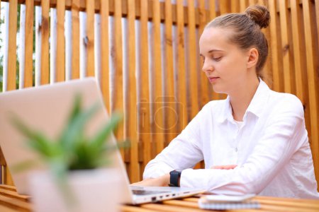 Concentrated woman wearing white shirt sitting in outdoor cafe working on laptop watching webinar online learning freelance job networking browsing.