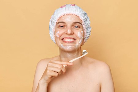 Foto de Mujer alegre sosteniendo cepillo de dientes haciendo procedimientos de higiene y belleza aplica crema usa gorra de ducha haciendo procedimientos de rutina de la mañana. - Imagen libre de derechos