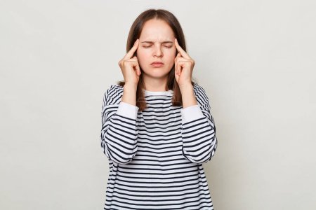 Photo for Sad unhealthy sick brown haired woman wearing striped shirt posing isolated over gray background having headache massaging her temples standing with closed eyes. - Royalty Free Image