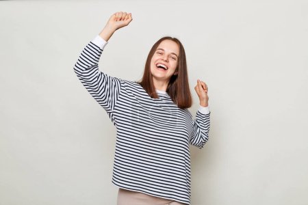 Photo for Extremely happy positive brown haired woman wearing striped shirt posing isolated over gray background standing with raised arms dancing celebrating holiday. - Royalty Free Image