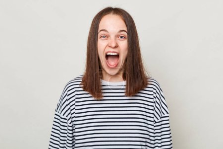 Photo for Crazy funny brown haired woman wearing striped shirt posing isolated over gray background standing with widely opened mouth screaming loud looking at camera. - Royalty Free Image