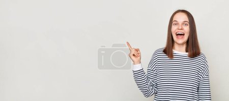 Photo for Amazed attractive funny brown haired woman wearing striped shirt posing isolated over gray background looking at camera with happy face pointing at advertisement area. - Royalty Free Image