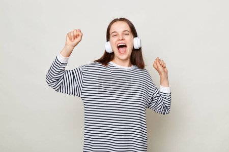 Photo for Positive happy cheerful brown haired woman wearing striped shirt posing isolated over gray background standing with headphones dancing with raised arms. - Royalty Free Image