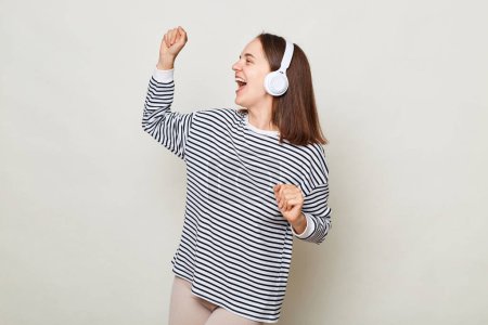 Photo for Extremely happy brown haired woman wearing striped shirt posing isolated over gray background being in good festive mood dancing listening to music in headphones. - Royalty Free Image
