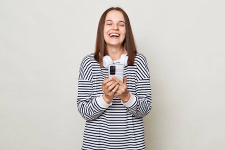 Photo for Happy positive brown haired woman wearing striped shirt posing isolated over gray background holding cell phone in hands chatting with friends checking social networks, - Royalty Free Image