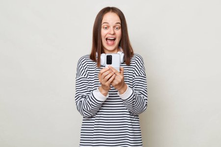 Photo for Amazed laughing brown haired woman wearing striped shirt posing isolated over gray background using mobile phone reading positive news looking at gadget display. - Royalty Free Image