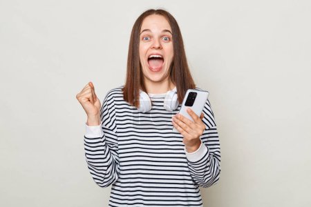 Photo for Extremely happy amazed brown haired woman wearing striped shirt posing isolated over gray background standing with clenched fist holding mobile phone gets sms. - Royalty Free Image