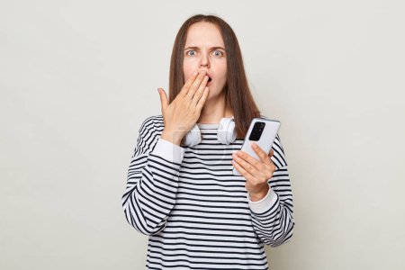Photo for Shocked surprised brown haired woman wearing striped shirt posing isolated over gray background holding smart phone covering mouth with palm astonished internet news. - Royalty Free Image