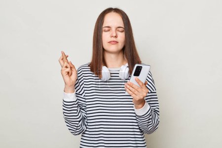 Photo for Hopeful brown haired woman wearing striped shirt posing isolated over gray background holding smart phone and crossed fingers waiting for important message. - Royalty Free Image