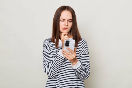 Photo for Pensive thoughtful brown haired woman wearing striped shirt posing isolated over gray background using cell phone, thinking what to answer on massage. - Royalty Free Image