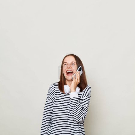 Photo for Laughing brown haired woman wearing striped shirt posing isolated over gray background talking on mobile phone copy space fore advertisement, mockup. - Royalty Free Image
