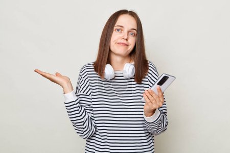 Photo for Confused puzzled brown haired woman wearing striped shirt posing isolated over gray background shrugging shoulders holding mobile phone. - Royalty Free Image