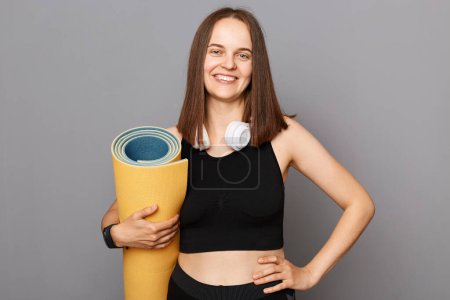 Photo for Health is wealth. Fitness starts with a workout. Find balance through yoga. Smiling woman standing carrying yoga mat isolated over gray background - Royalty Free Image