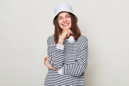 Photo for Cheerful woman wearing striped shirt and panama standing with headphones over neck isolated over gray background being in good mood smiling to camera holding chin. - Royalty Free Image