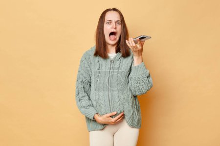 Photo for Despair shocked scared pregnant woman wearing knitted sweater standing isolated beige background holding smartphone recording voice messages calling doctor screaming feeling pain. - Royalty Free Image